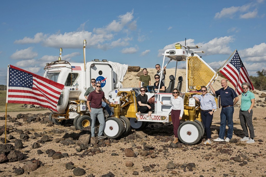 A group of people stand beside a large NASA rover vehicle in a rocky desert area, with American flags displayed.