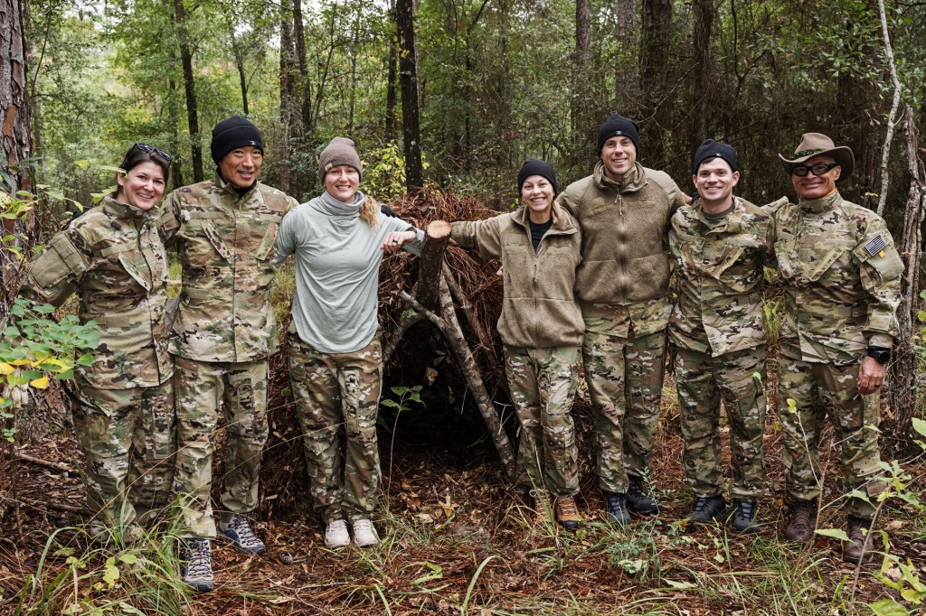 A group of people wearing camouflage uniforms stand together in a forest, smiling beside a small shelter made of branches.