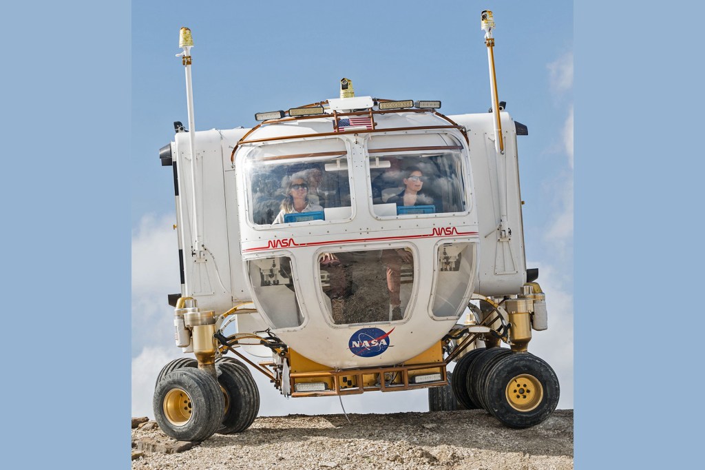 Two people sit inside a large NASA rover vehicle with wide wheels as it drives over rocky ground under a blue sky.