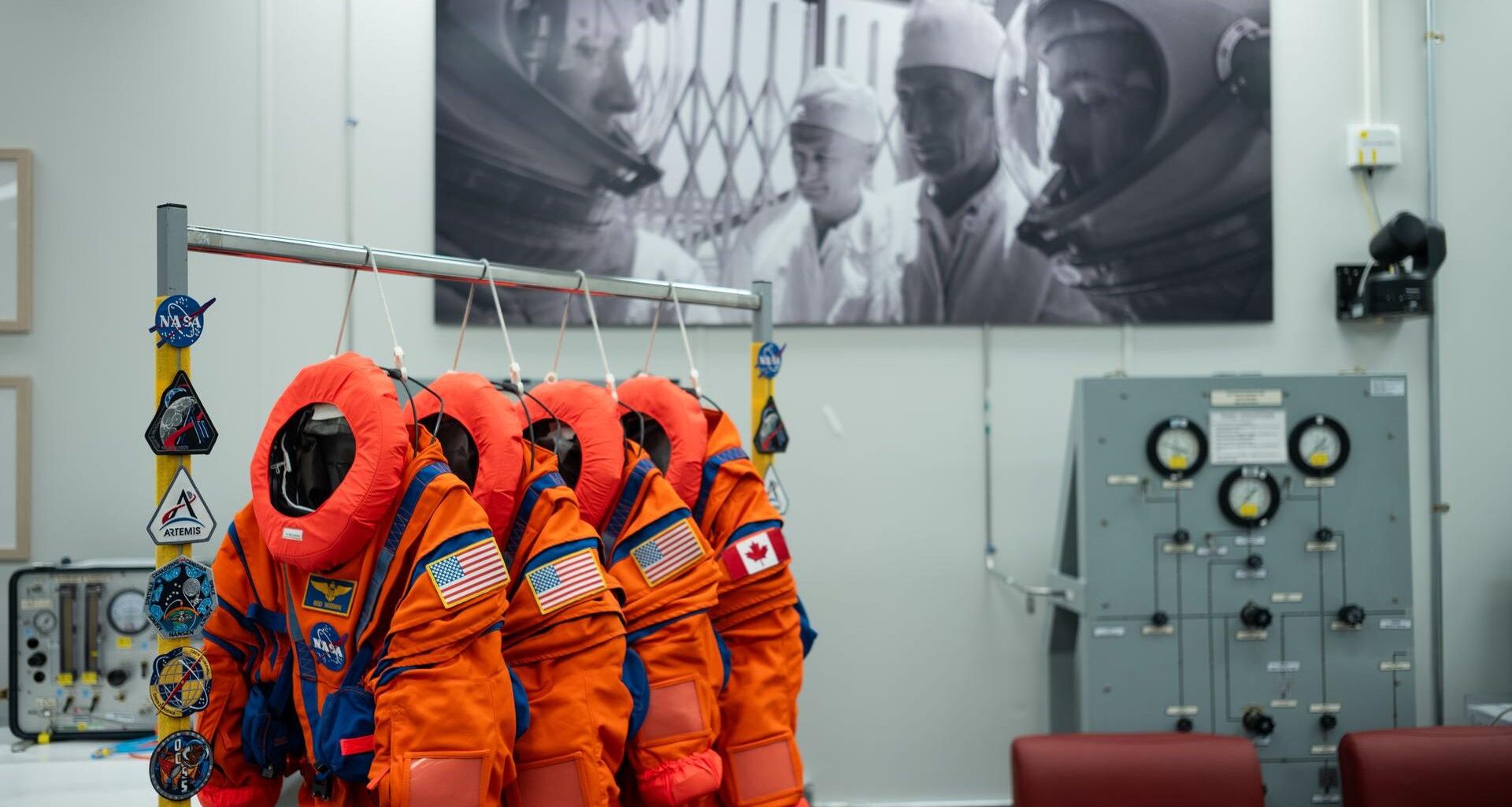 Four bright orange spacesuits with the circular NASA logo and American and Canadian flags on the arms hang on a rack in the middle of an empty room with chairs to the right and an image on the back white wall of two astronauts looking at each other