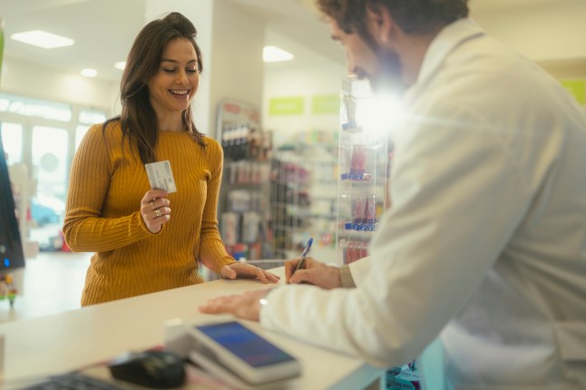 A woman stands at the pharmacy counter, smiling slightly while holding a credit, ready to complete her transaction