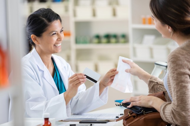 Pharmacist and customer interact at pharmacy counter, exchanging payment and prescription.