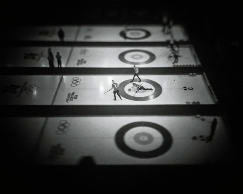 Black and white photo of several curling rinks viewed from above, with players standing on the ice near circular targets. The image has a dramatic, blurry vignette effect and Olympic rings are visible on the ice.