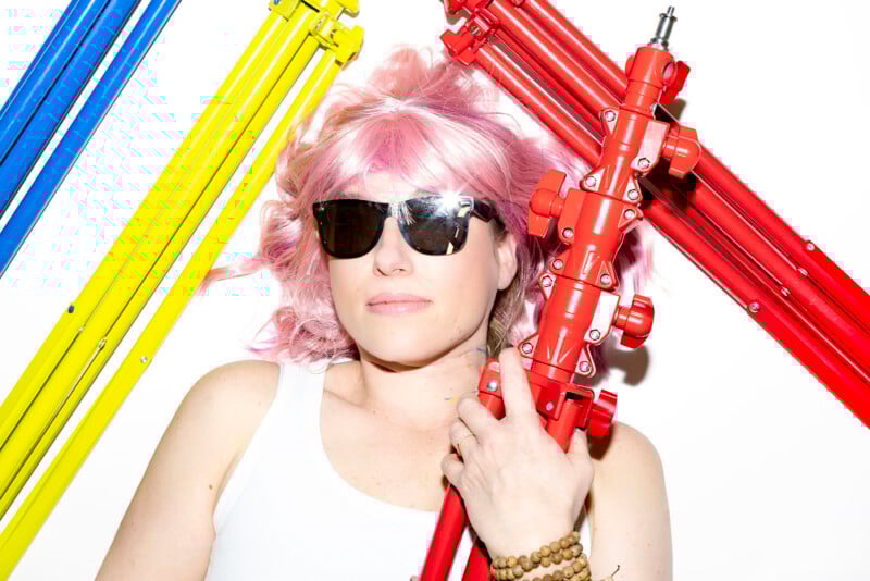 A person with pink hair and sunglasses lies on a white background, holding a red metal tripod. Yellow, blue, and red tripods are arranged around them. The person wears a white tank top and beaded bracelets.