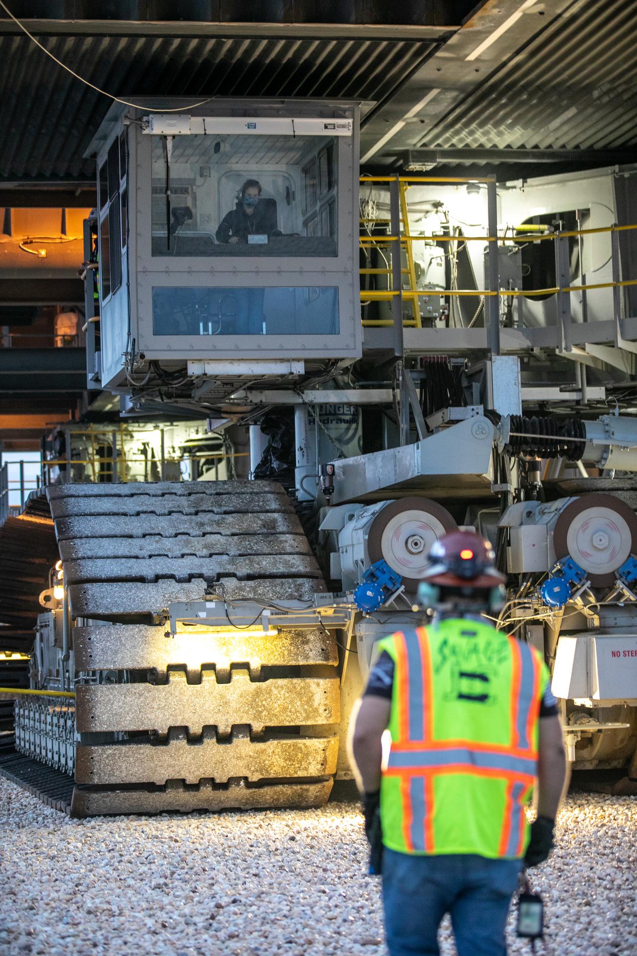 A woman drives NASA's crawler-transporter. She is visible inside the cab, seen through a large clear window. In the foreground, a man in a hardhat and high-visibility vest watches her from the ground.