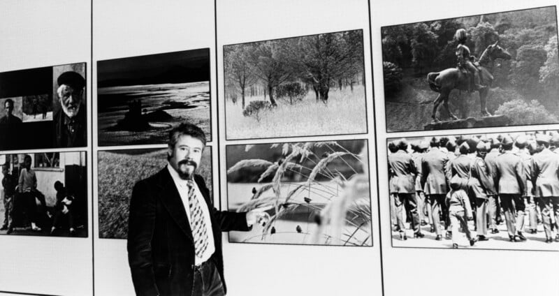 A man in a suit stands in front of a display wall featuring various black-and-white photographs, including landscapes, people, a statue on horseback, and birds on wire.