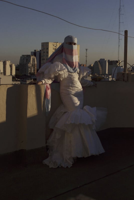 A person in a white ruffled gown and a pink, blue, and white face veil stands on a rooftop at sunset, with city buildings in the background and soft light casting long shadows.
