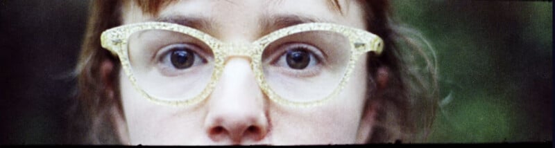 A close-up of a person’s face focusing on their eyes, wearing large, translucent, patterned eyeglasses. The background is blurry and green, drawing attention to the glasses and eyes.