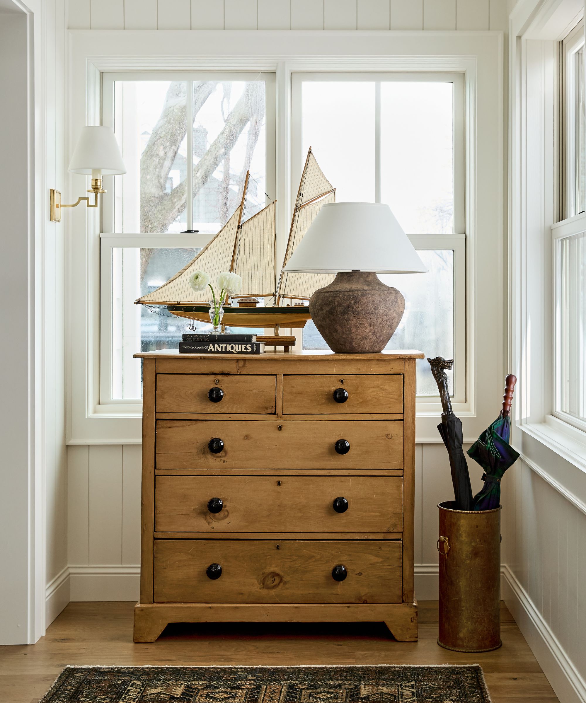 window vignette with a vintage wooden dresser styled with a vintage rug, antique boat, rustic lamp and books