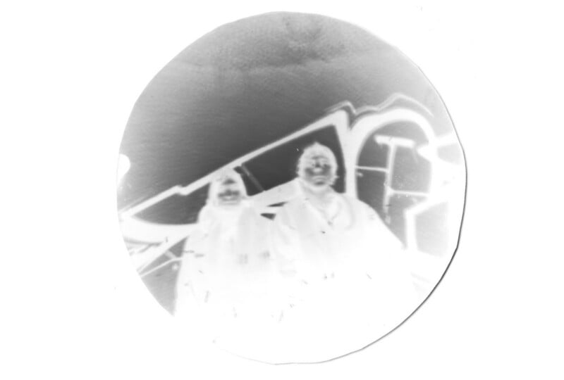 A black-and-white circular photo negative shows two people standing in front of a wall with graffiti. Their facial features are faint, and the image has a high-contrast, inverted look.