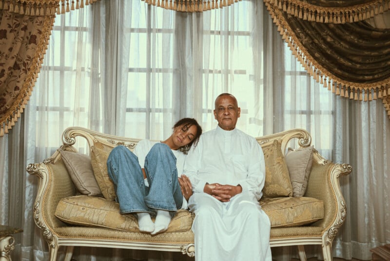 A young woman leans her head on an older man's shoulder as they sit on an ornate sofa in a room with large windows and luxurious drapes. The man wears a white robe; the woman is in jeans and a sweater, both looking content.