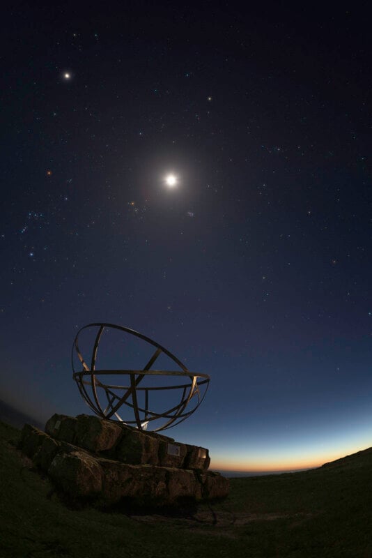 A metal sundial sits on stone blocks under a night sky filled with stars and a bright moon, with a faint glow from the setting or rising sun on the horizon.