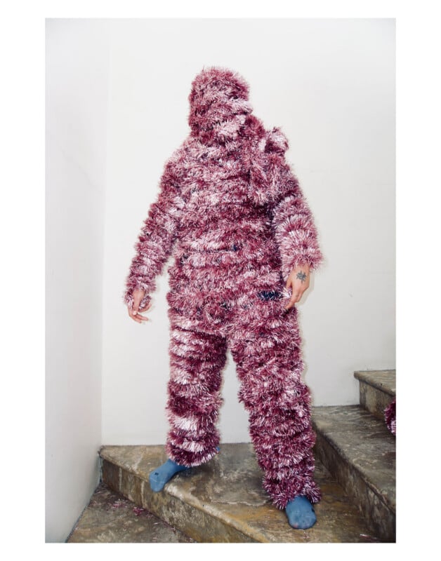A person stands on stairs wearing a full-body suit made of pink and silver tinsel, obscuring their face and features. Their hands are exposed, and they are wearing blue slippers. The background is a plain white wall.