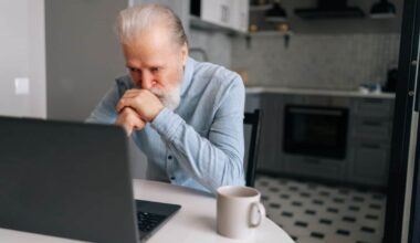 Portrait of pensive bearded senior looking on screen of laptop sitting at table with coffee cup.