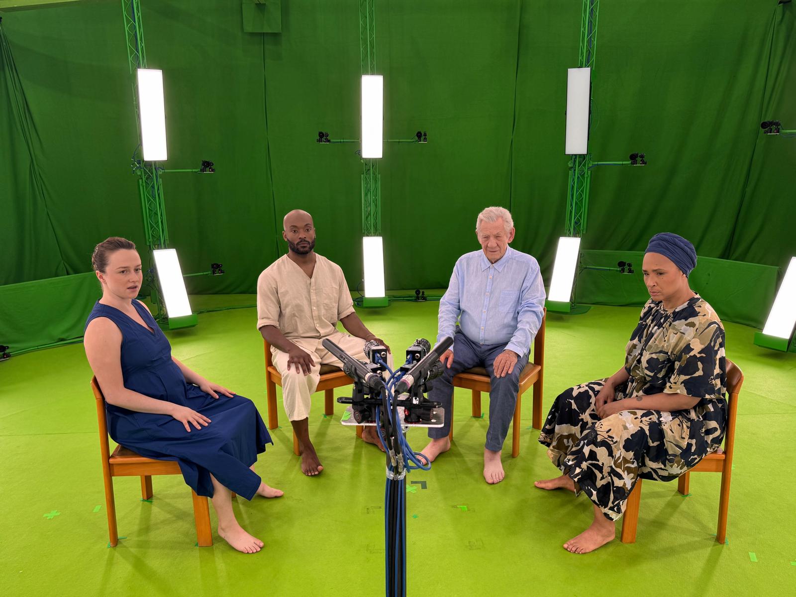 Two men and two women sitting in a semi circle on chairs. The room around the people is covered in green fabric, the floor is also green, and the group are all looking into an array of cameras.