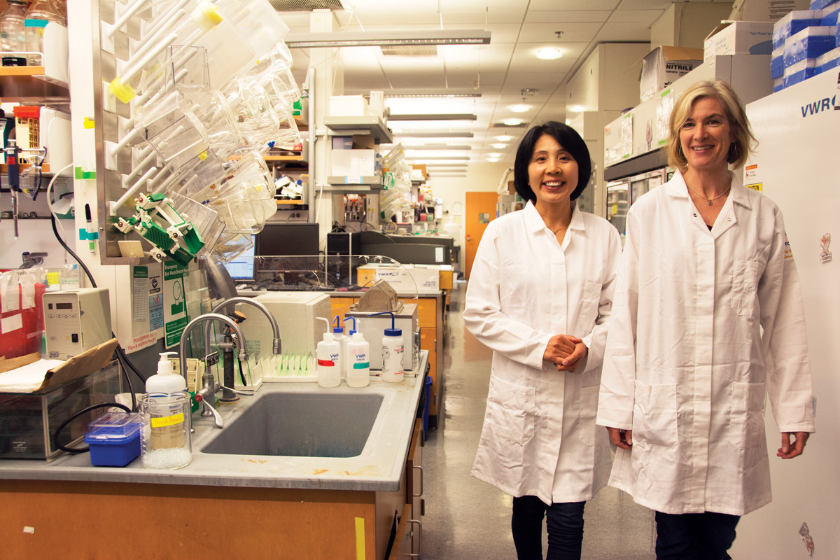 Two women wearing white lab coats stand in a brightly lit chemistry lab with a sink, a drying rack, and shelves full of equipment around them.