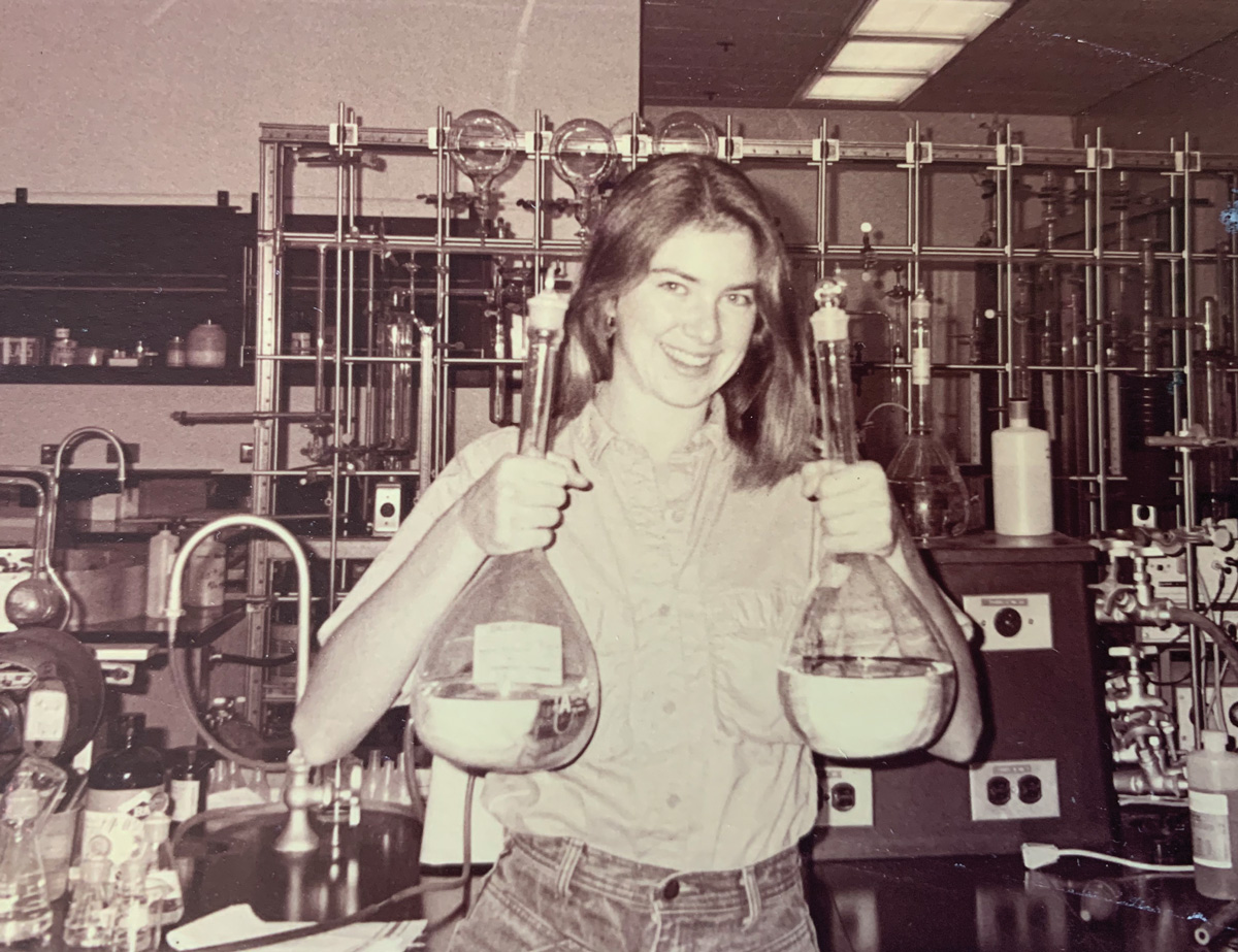 A black-and-white photo of a young woman holding two flasks filled with clear liquid and standing in front of a lab bench filled with equipment.