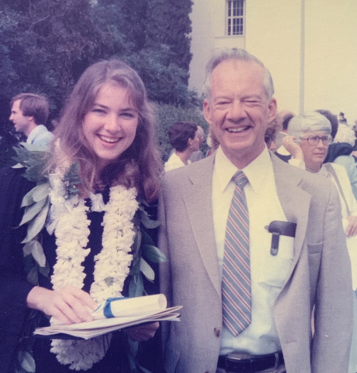 A young woman wearing a lei and holding a diploma stands next to a man wearing a suit jacket and tie, as both smile.