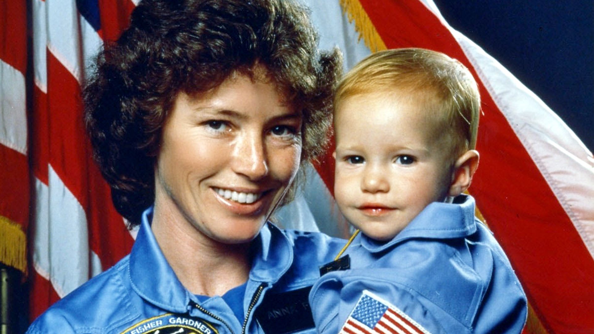 Astronaut Anna Fisher with her baby, stood in front of the American flag.