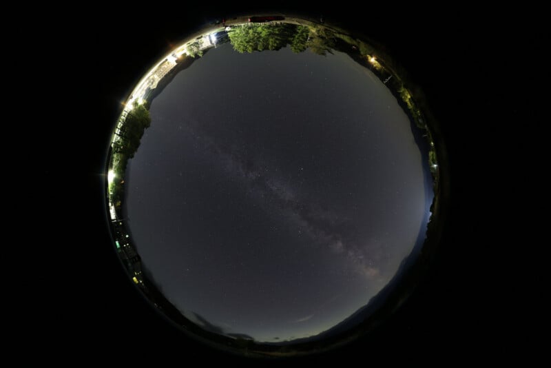 A fisheye lens photo shows the night sky filled with stars and the Milky Way, surrounded by a circular border of trees, mountains, and faint lights from a small settlement.