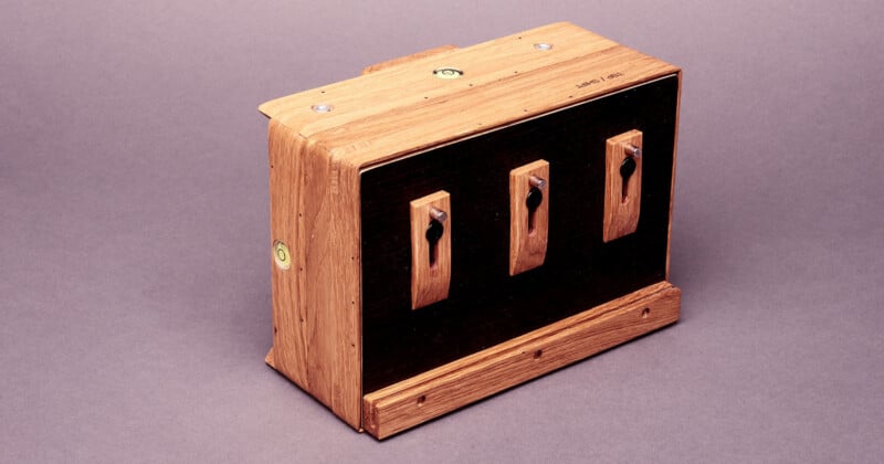 A wooden box with three vintage-style toggle switches mounted on a black panel. The box has a natural wood finish and metal fasteners, and is set against a plain, light purple background.