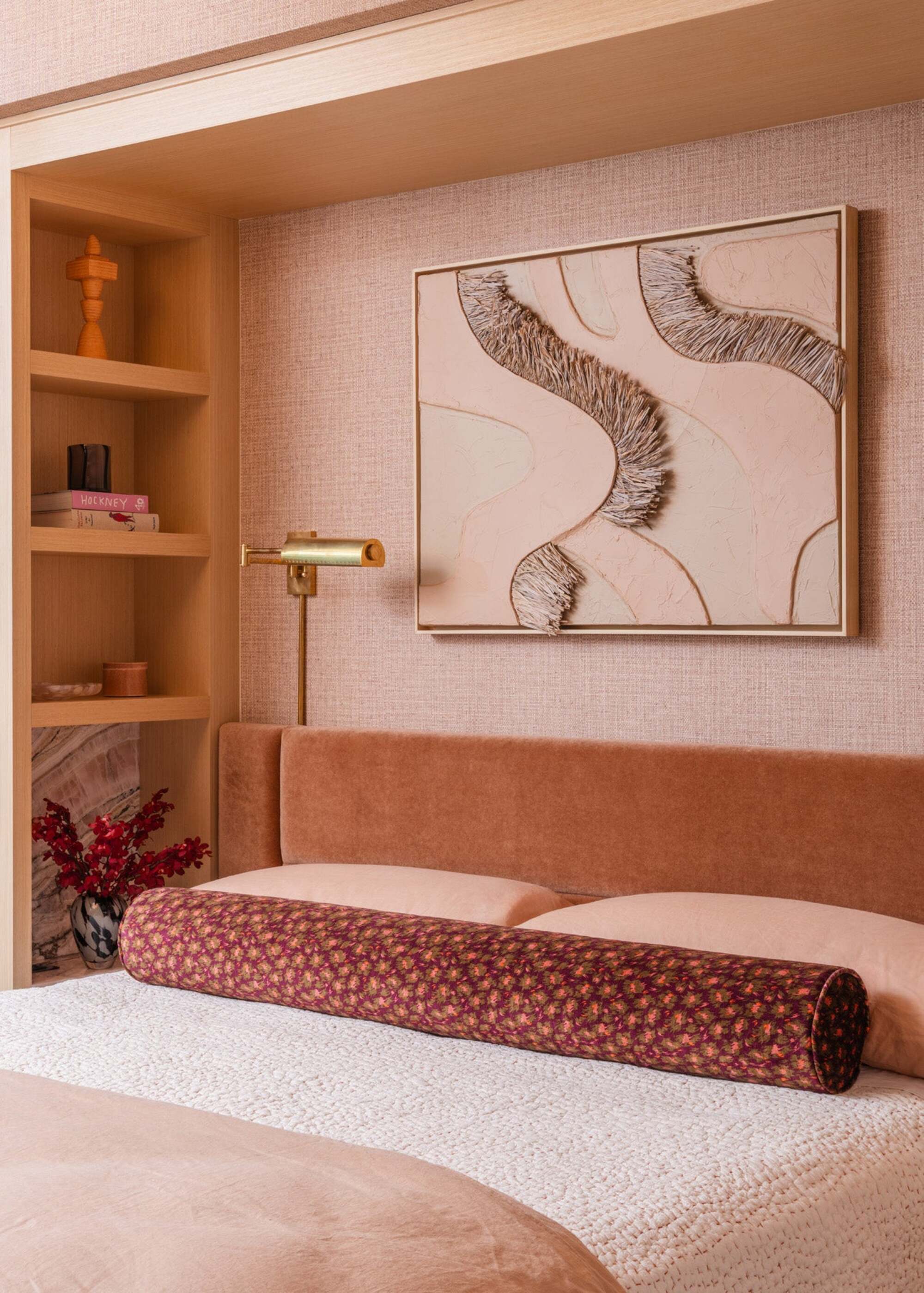 bedroom with extended velvet coral headboard, peach sheets with white waffle blanket, patterned red bolster cushion, built-in shelves with grasscloth wallpaper and large artwork