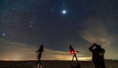 A man looks up at the moon glowing in a starry sky in a field next to two telescopes. A glow lights up clouds near the horizon.
