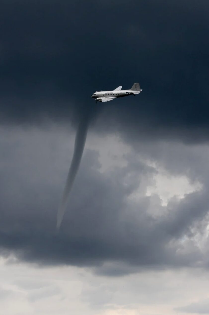 A plane flies over a long funnel cloud in grey weather