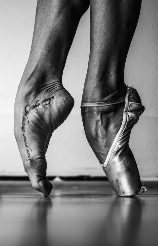 Black and white close-up of a ballet dancer’s feet en pointe; one foot is bare with a tattoo on the arch, and the other wears a worn ballet shoe, both poised gracefully on a smooth floor.