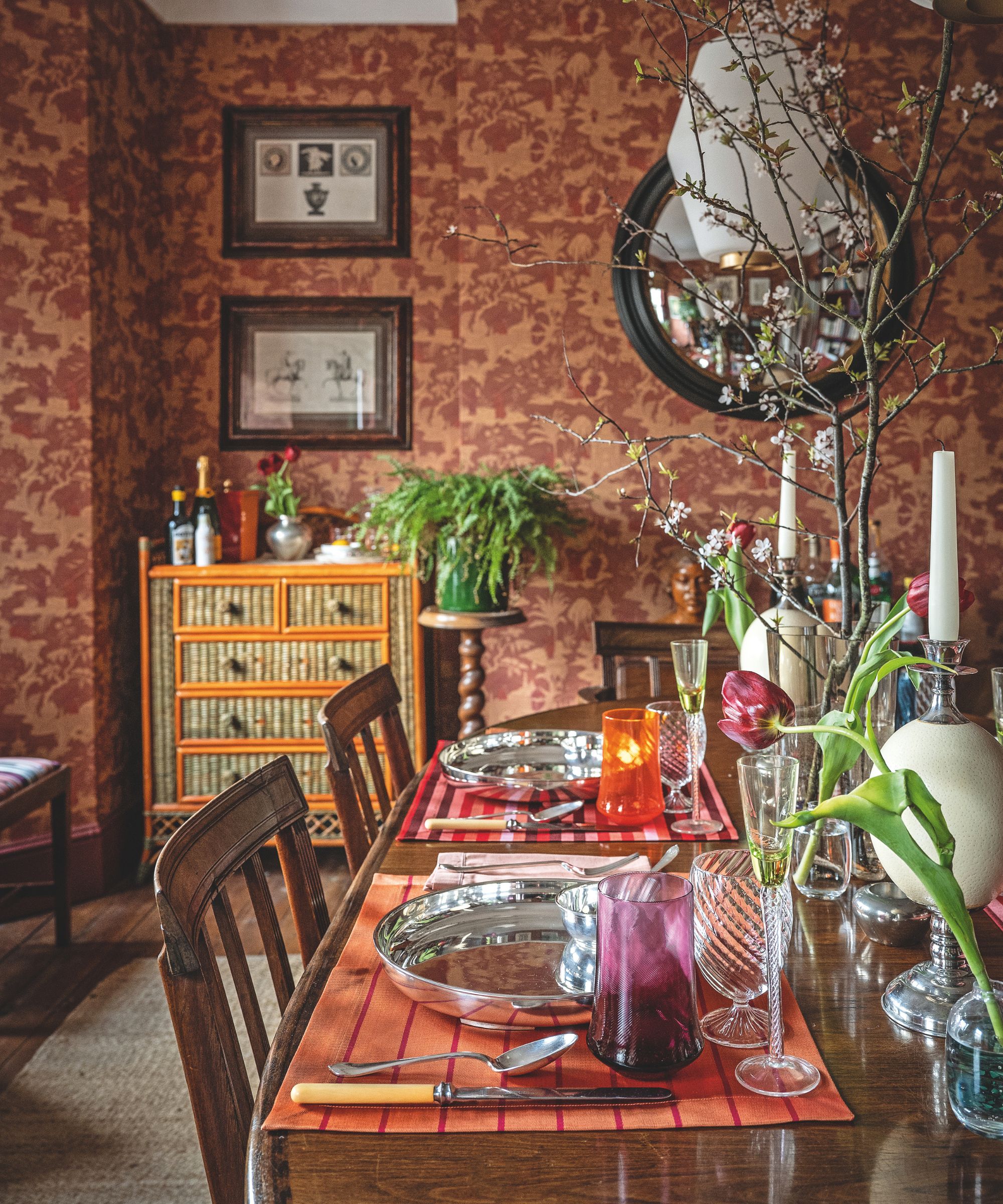 A moody dining room with red patterned wallpaper and a wooden table set with silver plates, colorful glassware, and a centerpiece of flowering branches