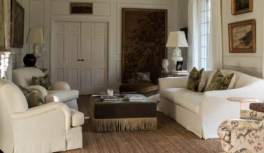 a traditional white living room with a large antiqu tapestry and vintage art, wall moldings, two white armchairs, a linen couch, green velvet fringed ottoman