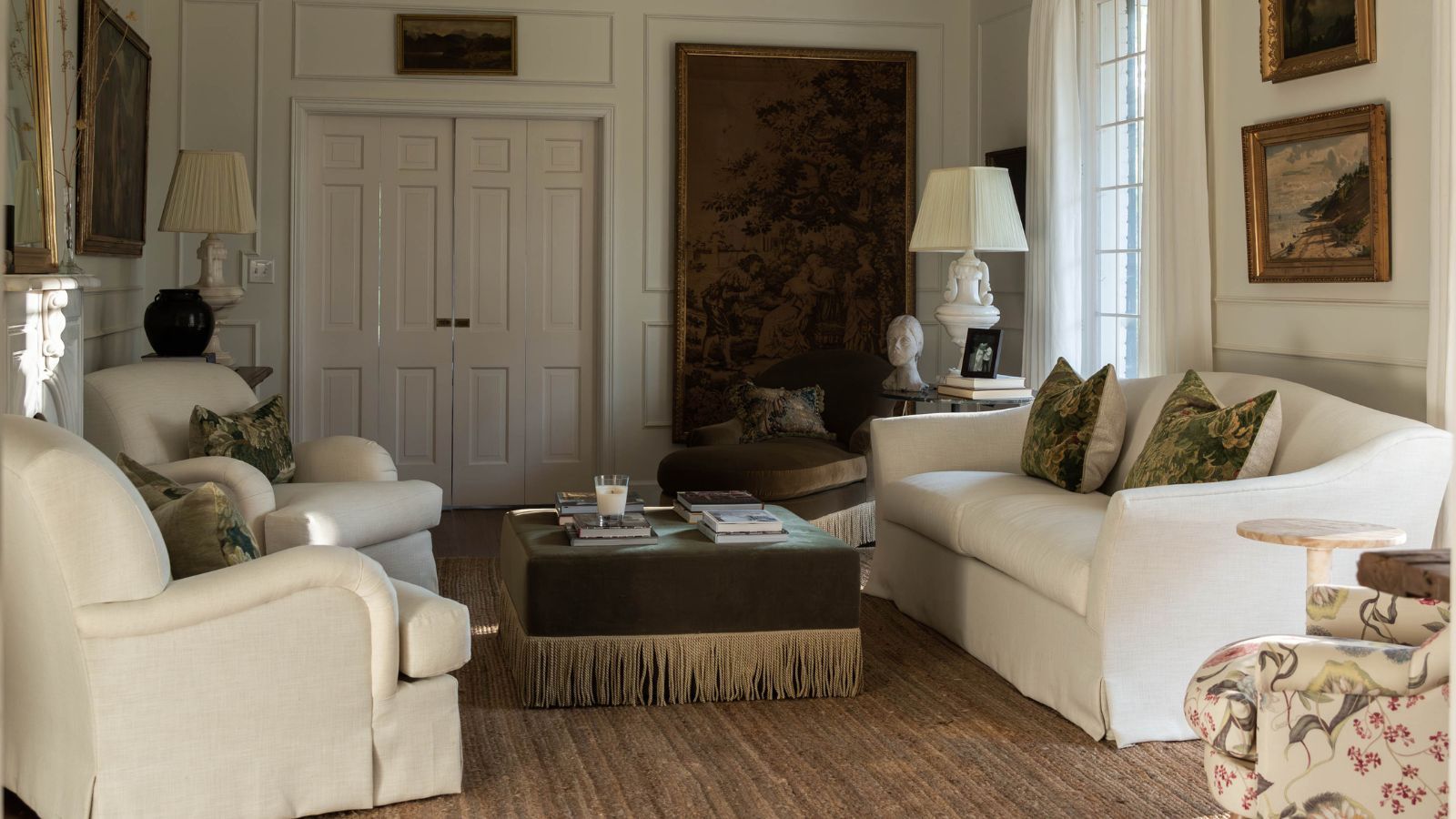 a traditional white living room with a large antiqu tapestry and vintage art, wall moldings, two white armchairs, a linen couch, green velvet fringed ottoman
