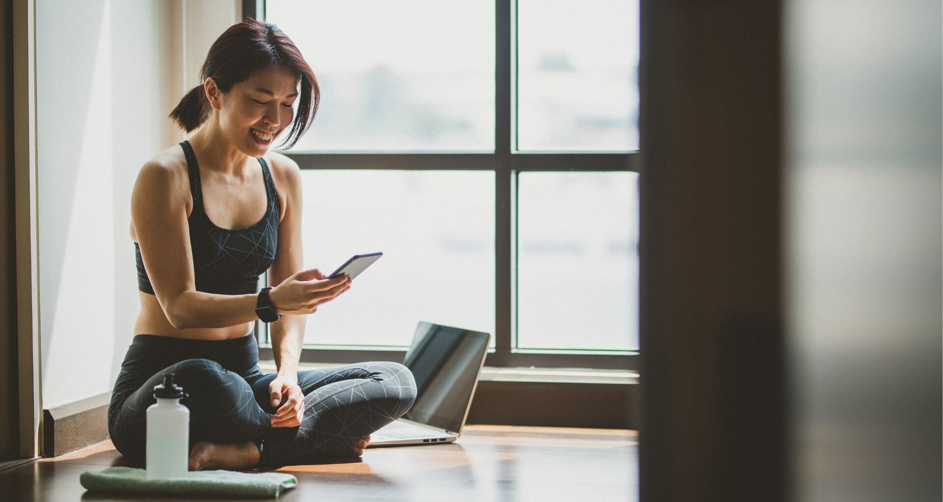 Woman looking down at phone smiling with water bottle and laptop