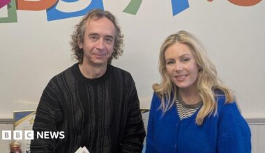 Geoff Russell-Jones in a black jumper with curly hair smiles at the camera while standing next to Rebecca Caswell-Fox, who has long blonde hair and a blue cardigan.
