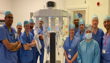 A group of people dressed in blue hospital scrubs, standing around a white piece of robotic equipment. They are standing in a light-coloured room, which appears to be a hospital setting.