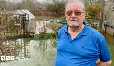 A man in a blue polo shirt and dark glasses is standing in a flooded domestic garden. Bushes, plants, a garage, greenhouse and washing pole is partially submerged. There is a wood panelled fence around the garden. Trees stand behind the fence in a field which is also flooded.
