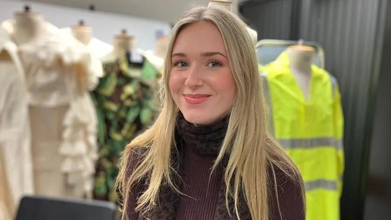 Hollie Ralph has long blonde hair and blue eyes and wears a brown polo neck jumper and a patterned scarf. Behind her are several mannequins displaying different outfits, including a cream-colored ruffled garment, a camouflage-patterned outfit with leaf-like details and bright yellow high‑visibility jacket.
