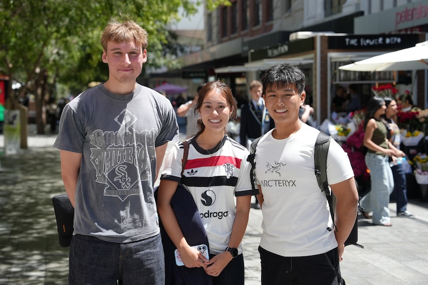Three young people, two men and a woman, stand together outside and smile.
