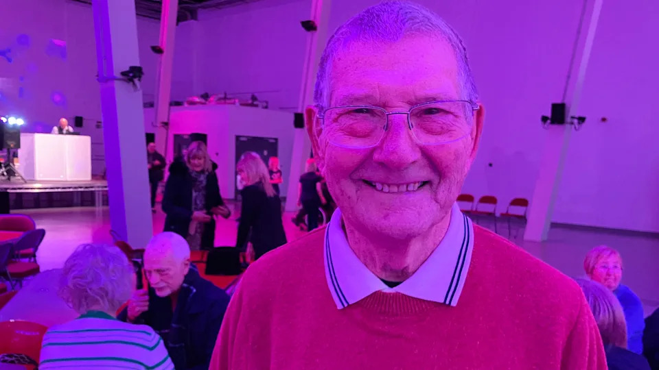 A man with grey hair and glasses, in a red jumper and white polo shirt, at a hall which has pink lights and is being used as the venue for a disco for older people