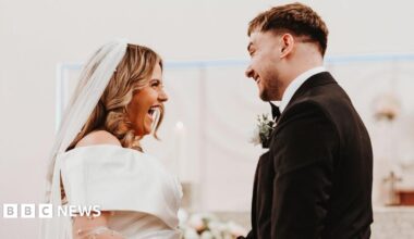 Caoimhe is smiling at Ollie. They are at the alter of their wedding. She is wearing a white veil and an off the shoulder wedding dress. He is wearing a dark coloured suit and has short dark hair. She has long blonde hair curled.