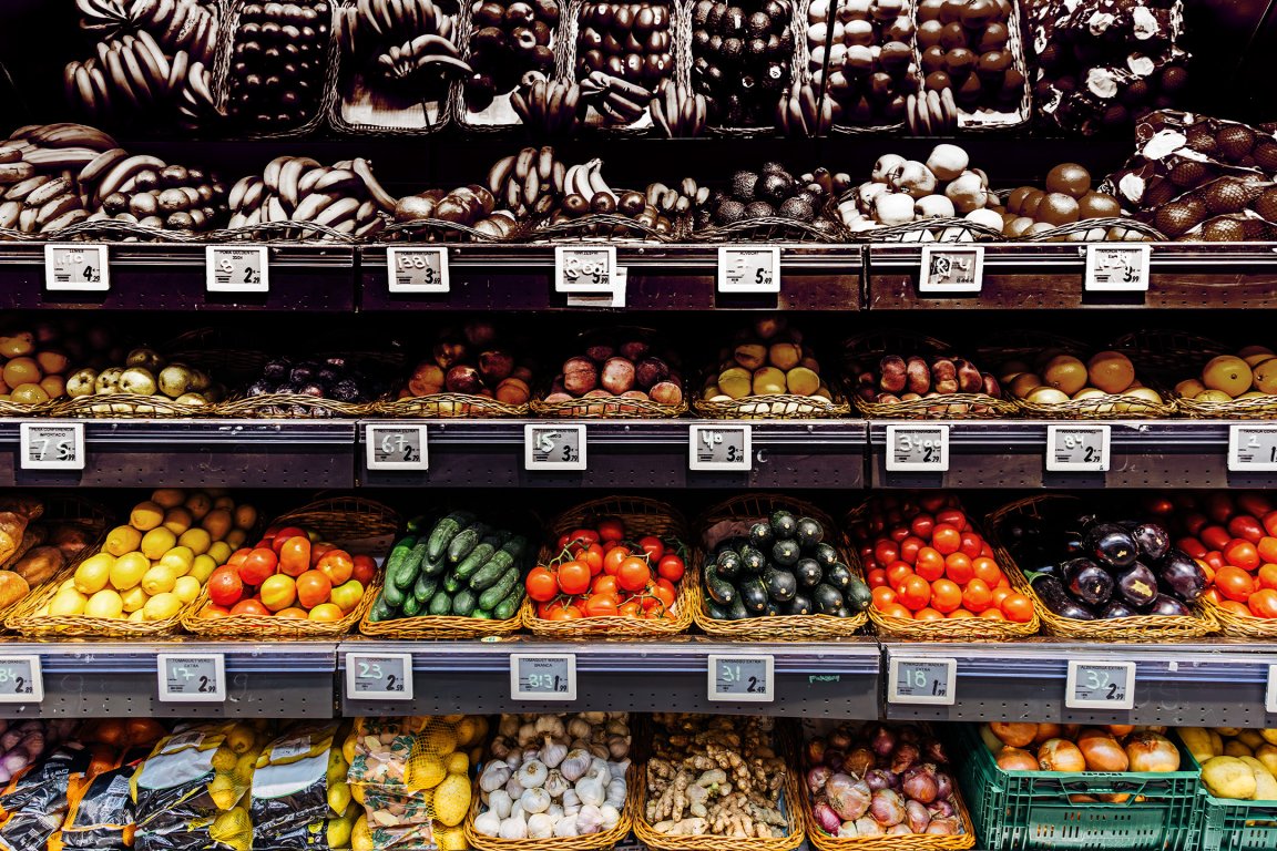 Shelves filled with a variety of fresh fruits and vegetables arranged in baskets, including bananas, tomatoes, cucumbers, lemons, eggplants, garlic, ginger, and onions, with price tags displayed below each section.