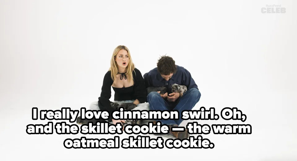 Two people sit on the floor with several puppies. The person on the left wears a black top; the person on the right, a dark shirt and jeans