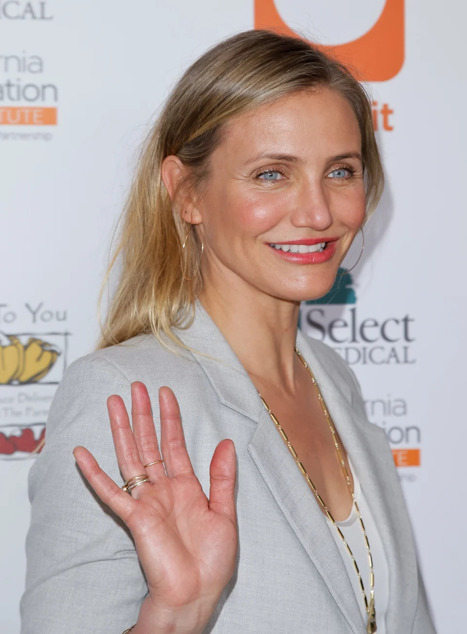 A person waves and smiles at an event, wearing a blazer over a shirt with thin necklaces