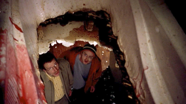 Jesse Pinkman, wearing a beanie and red sweater, and Walter White, wearing a yellow shirt and jacket, look up at the hole in their ceiling leading to bloody bathtub in 