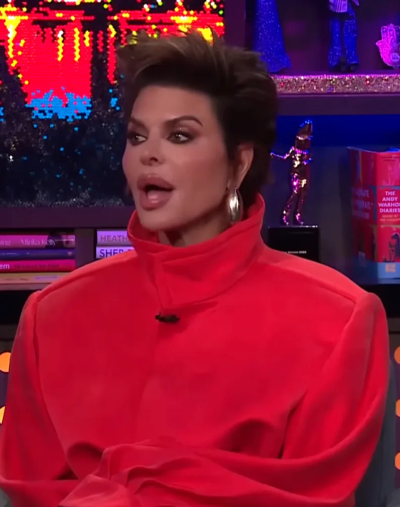 Person in a high-neck red outfit speaking during a talk show, seated in front of bookshelves