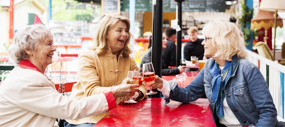 Three older woman sit on a patio, clinking together glasses of wine.