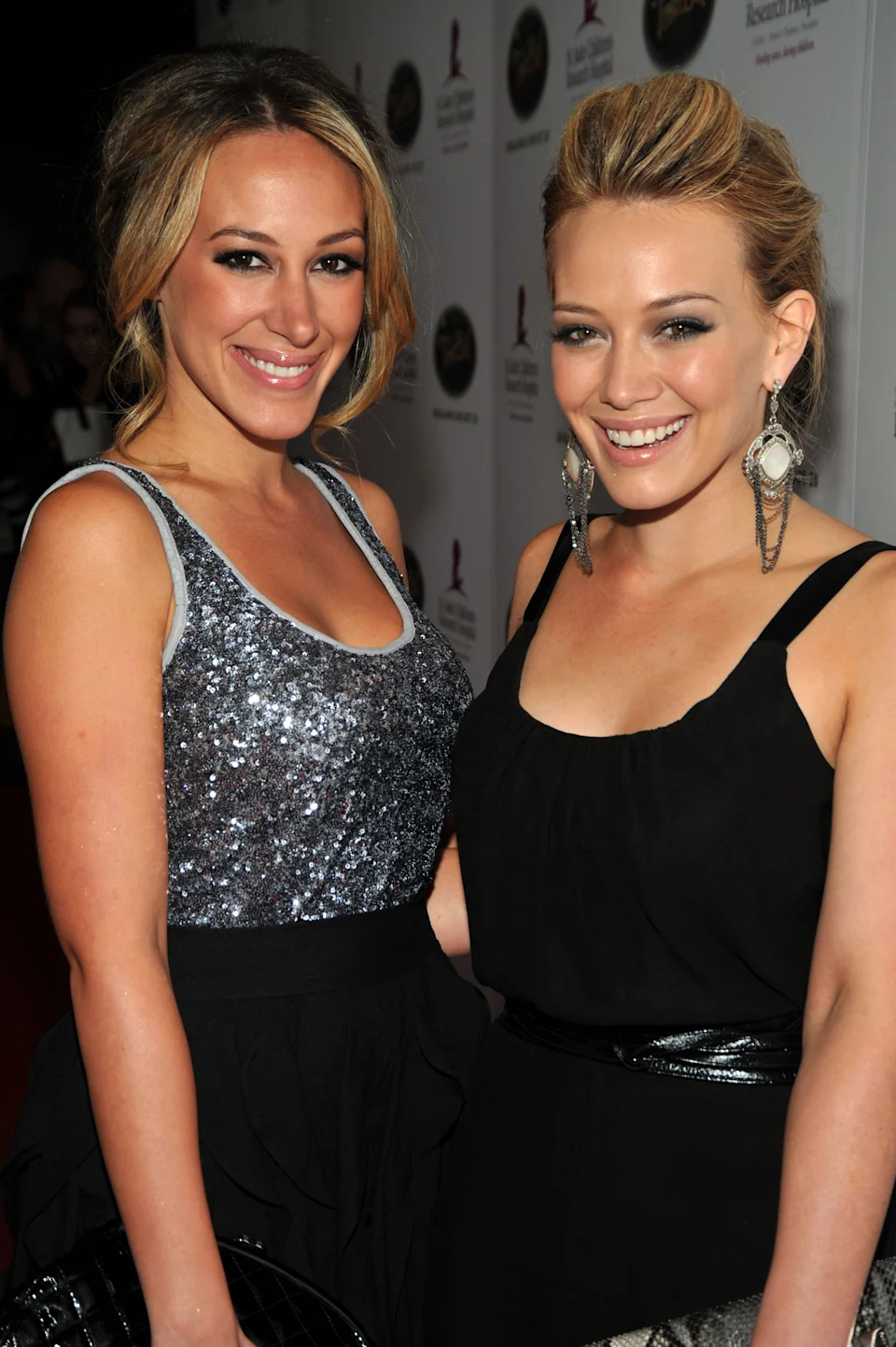 Two people smiling on the red carpet. One wears a sparkly top; the other, a black dress with bold earrings