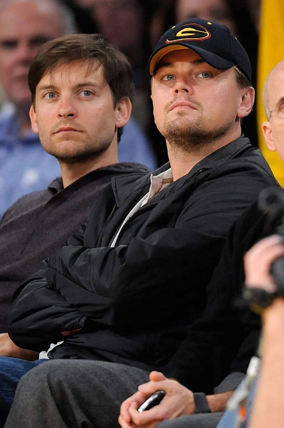 Two people in casual attire watch a sports event. One wears a baseball cap. They seem focused on the game