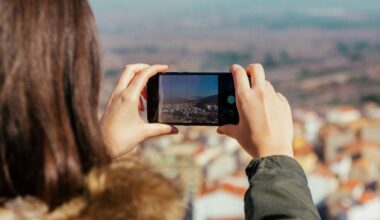 A person with long brown hair wearing a green jacket takes a photo of a scenic, sunlit landscape with their smartphone, capturing a distant town and natural surroundings.