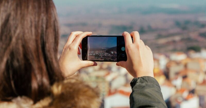 A person with long brown hair wearing a green jacket takes a photo of a scenic, sunlit landscape with their smartphone, capturing a distant town and natural surroundings.